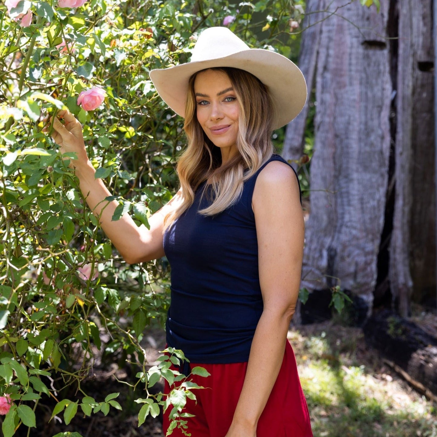 woman in hat in a garden with roses wearing a merino singlet in navy